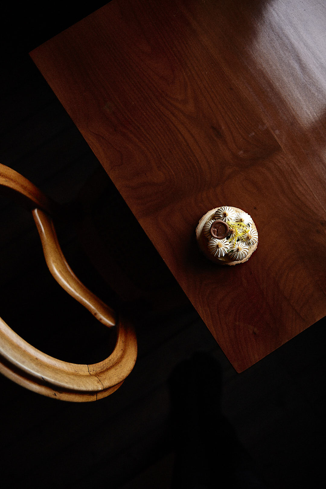 Produktfoto von einem Zitronen-Törtchen von Tartes&Törtchen aus Bielefeld, fotografiert auf einem Holztisch schräg im Anschitt, die Stuhllehne den am Tisch stehenden Holzstuhles ist sichtbar, der Boden liegt im Dunkeln