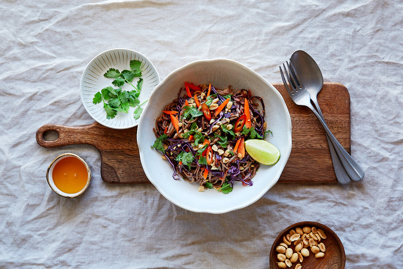 Food Foto von Thai-Nudel-Salat in weißer Schüssel auf Holzbrett mit Besteck und einzelnen Zutaten daneben